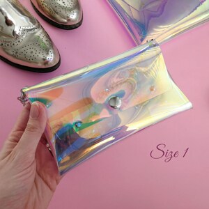 May include: A iridescent clear plastic clutch purse with a silver snap closure. The purse is on a pink surface with a watch and shoes in the background. The text "Size 1" is written on the surface.