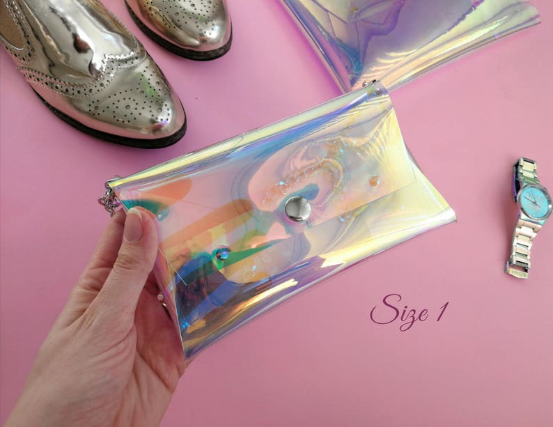 May include: A iridescent clear plastic clutch purse with a silver snap closure. The purse is on a pink surface with a watch and shoes in the background. The text "Size 1" is written on the surface.
