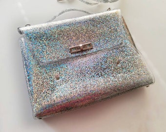Pode incluir: Uma bolsa de mão transparente de plástico prateado iridescente com um fecho de metal prateado.