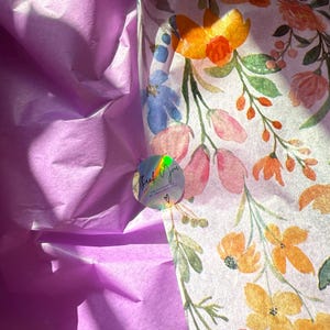 May include: A package presentation featuring purple tissue paper and floral wrapping paper. The wrapping paper has a white background with orange, pink, and blue flowers. A holographic sticker with the words "Thank You" is also visible.