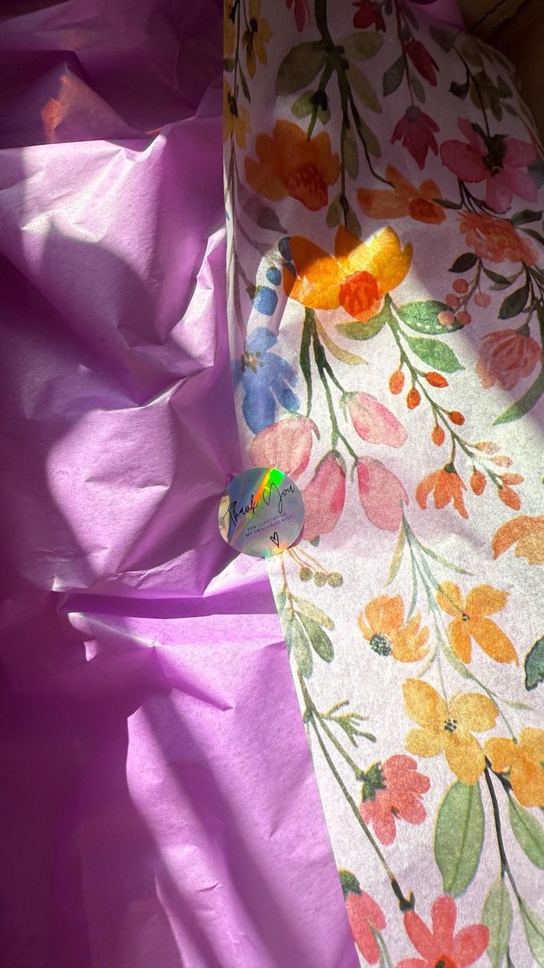 May include: A package presentation featuring purple tissue paper and floral wrapping paper. The wrapping paper has a white background with orange, pink, and blue flowers. A holographic sticker with the words "Thank You" is also visible.