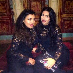 May include: Two women wearing black lace dresses with long sleeves. They are standing in front of a wooden wall with a distressed finish.