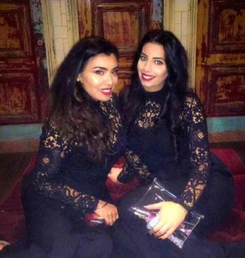 May include: Two women wearing black lace dresses with long sleeves. They are standing in front of a wooden wall with a distressed finish.