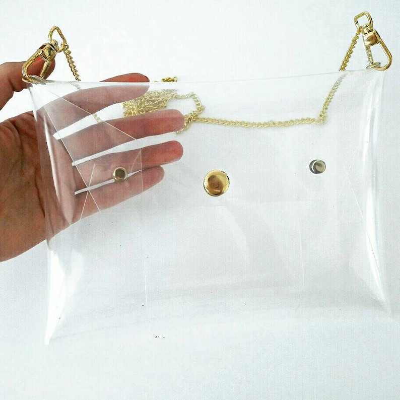 May include: A clear plastic clutch bag with a gold chain strap and gold-toned hardware. The bag has a flap closure with a snap button.