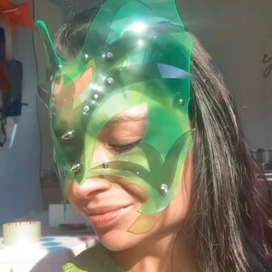 Op de afbeelding: Een groen plastic masker met een geometrisch ontwerp en zilveren studs. Het masker bedekt de ogen en een deel van de neus en het voorhoofd.