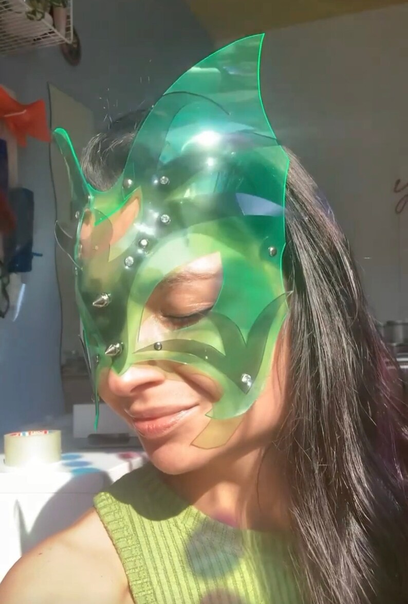 Op de afbeelding: Een groen plastic masker met een geometrisch ontwerp en zilveren studs. Het masker bedekt de ogen en een deel van de neus en het voorhoofd.