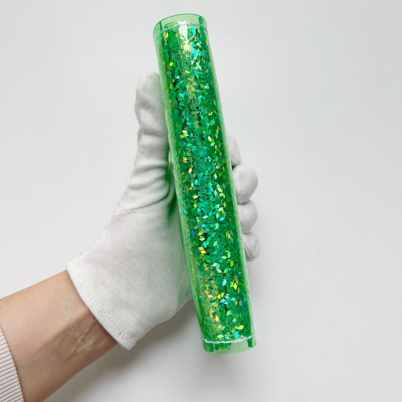 May include: A clear, cylindrical container filled with sparkling green glitter. The glitter is composed of small, diamond-shaped pieces. The container is held by a gloved hand against a white background. The container is approximately 10 inches long.