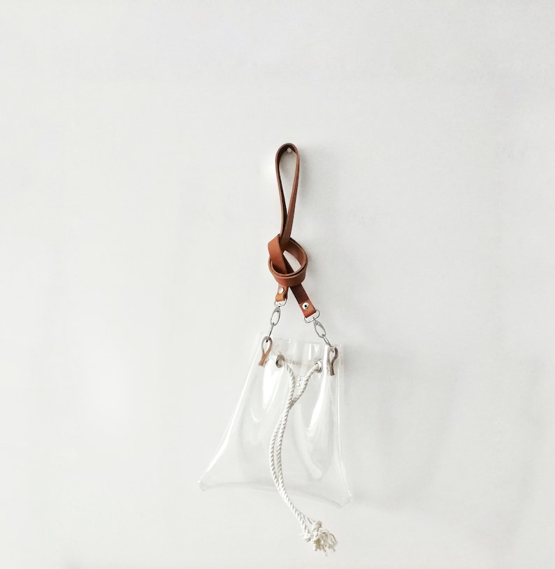 May include: A clear transparent pouch with a white rope drawstring closure. The pouch features silver-toned metal hardware connecting to a brown leather wrist strap that is tied into a knot at the top, providing a functional carrying handle.