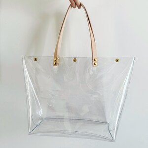 May include: A clear tote bag with light tan leather handles and gold-colored rivets. The bag is made of transparent material, allowing visibility of the contents. The handles are attached to the bag with gold rivets.