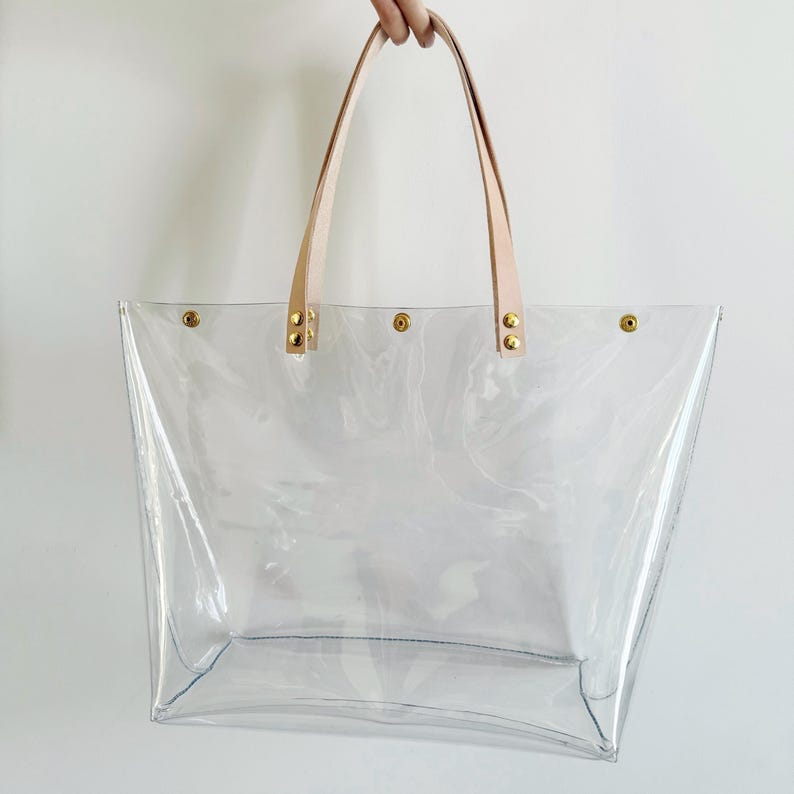 May include: A clear tote bag with light tan leather handles and gold-colored rivets. The bag is made of transparent material, allowing visibility of the contents. The handles are attached to the bag with gold rivets.