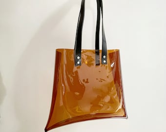 May include: A clear, amber-colored tote bag with black leather handles. The bag has a rectangular shape with a unique, flared design on the sides. The handles are attached with silver rivets.