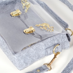 May include: A gray felt handbag with a clear plastic front panel. The bag features gold-colored hardware, including clasps, decorative filigree, and a shoulder strap with a gold clasp. The bag has a rectangular shape.
