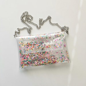 May include: A clear, rectangular crossbody bag filled with colorful confetti. The bag has a silver chain strap and a snap closure. The confetti includes various shapes and colors, creating a playful and festive look. The bag is made of a transparent material.