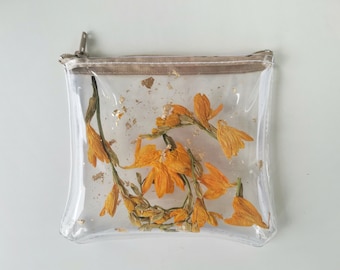 Puede incluir: Bolsa cuadrada transparente con cierre de cremallera, llena de flores naranjas secas y hojuelas doradas. La bolsa tiene un ribete beige y una cremallera plateada. Las flores son el punto focal, creando un accesorio artístico único.