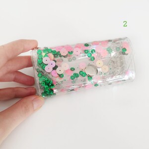May include: A clear plastic pouch with a silver button closure. The pouch is decorated with green and pink sequins.