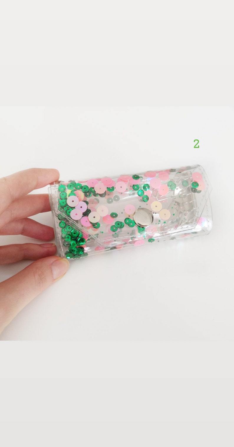 May include: A clear plastic pouch with a silver button closure. The pouch is decorated with green and pink sequins.