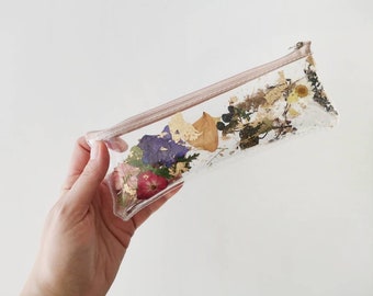 Puede incluir: Estuche rectangular transparente lleno de flores secas y pan de oro. El estuche tiene una cremallera y un ribete rosa claro. Las flores son de varios colores, incluyendo rojo, morado y amarillo. El estuche se sostiene en una mano.