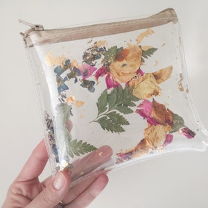 May include: Clear plastic zippered pouch with dried flowers and gold leaf accents. The flowers are pink, yellow, and blue.