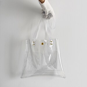 K&ouml;nnte beinhalten: Transparente Tragetasche mit rechteckiger Form und kleiner Fronttasche. Die Tasche hat zwei Griffe und wird von einer behandschuhten Hand gehalten. Die Tasche besteht aus transparentem Kunststoff und hat goldfarbene Nieten und einen goldfarbenen Druckknopfverschluss.