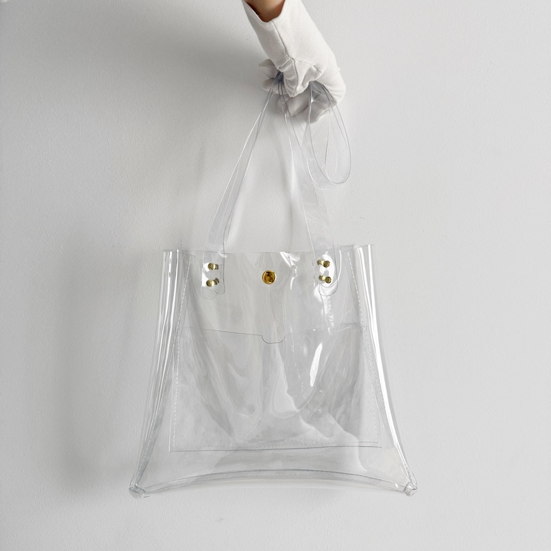 K&ouml;nnte beinhalten: Transparente Tragetasche mit rechteckiger Form und kleiner Fronttasche. Die Tasche hat zwei Griffe und wird von einer behandschuhten Hand gehalten. Die Tasche besteht aus transparentem Kunststoff und hat goldfarbene Nieten und einen goldfarbenen Druckknopfverschluss.