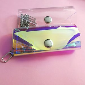 May include: A clear, iridescent key case featuring a metallic snap button closure and a silver-toned internal key holder with six hooks. The material displays a reflective, rainbow-like sheen, and the structure is rectangular with rounded edges.