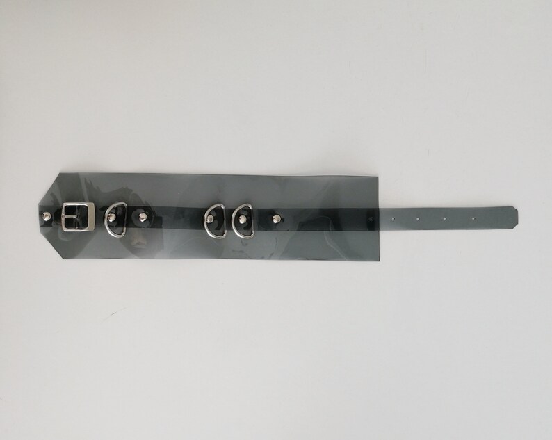 May include: A black leather choker with silver hardware, including a buckle, two D-rings, and three studs. The choker is transparent and has a black strap.