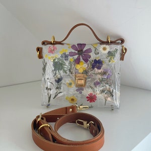 May include: A clear plastic handbag with a brown leather strap and handle. The bag is decorated with pressed flowers in shades of purple, yellow, and pink.