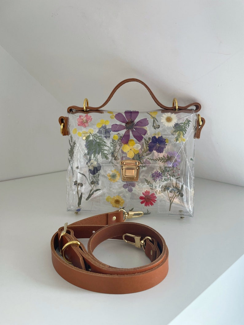 May include: A clear plastic handbag with a brown leather strap and handle. The bag is decorated with pressed flowers in shades of purple, yellow, and pink.