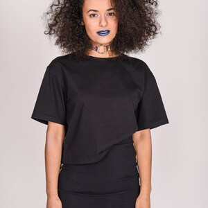 May include: A woman with curly dark hair wearing a black cropped t-shirt and a black skirt. She is wearing a colorful choker necklace and blue lipstick.