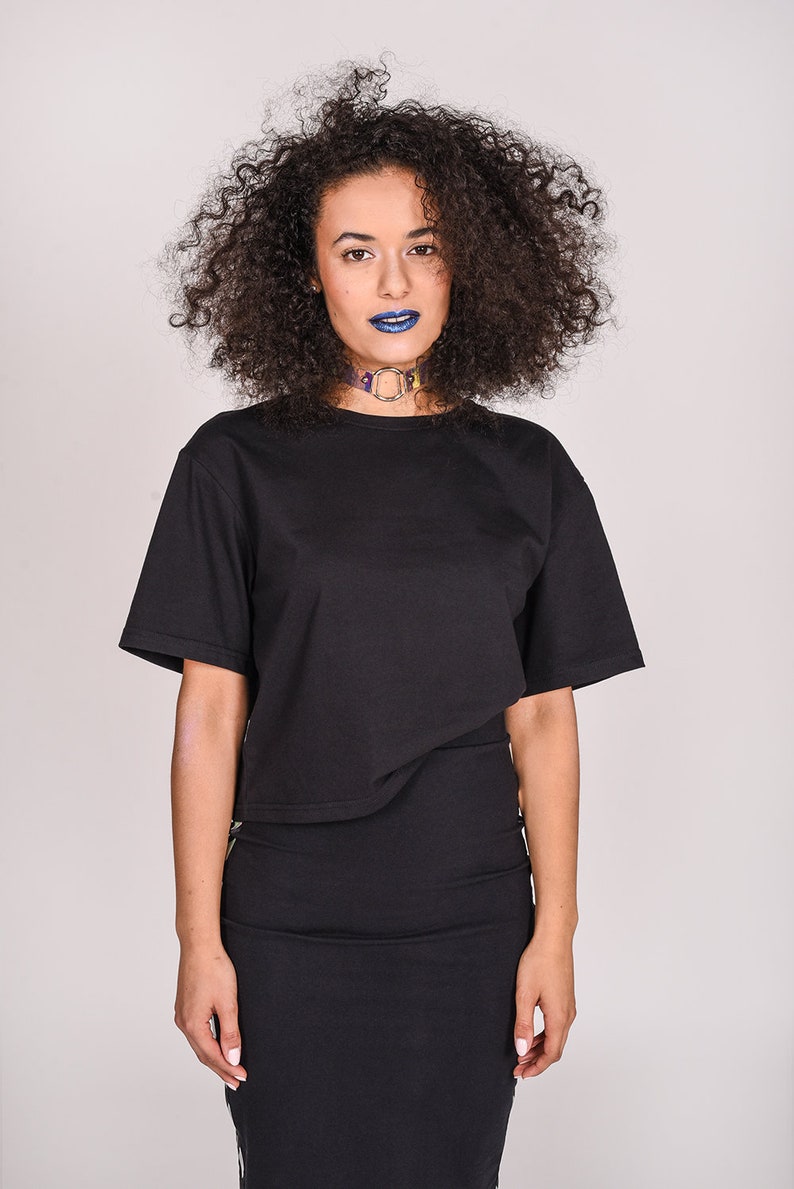 May include: A woman with curly dark hair wearing a black cropped t-shirt and a black skirt. She is wearing a colorful choker necklace and blue lipstick.