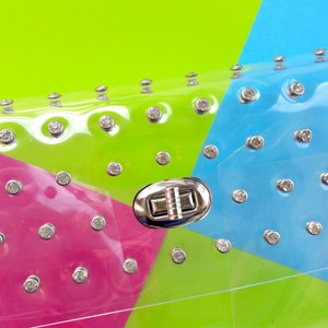 Puede incluir: Un bolso de mano transparente de pl&aacute;stico con un cierre plateado y pedrer&iacute;a. El bolso es rosa, verde y azul.
