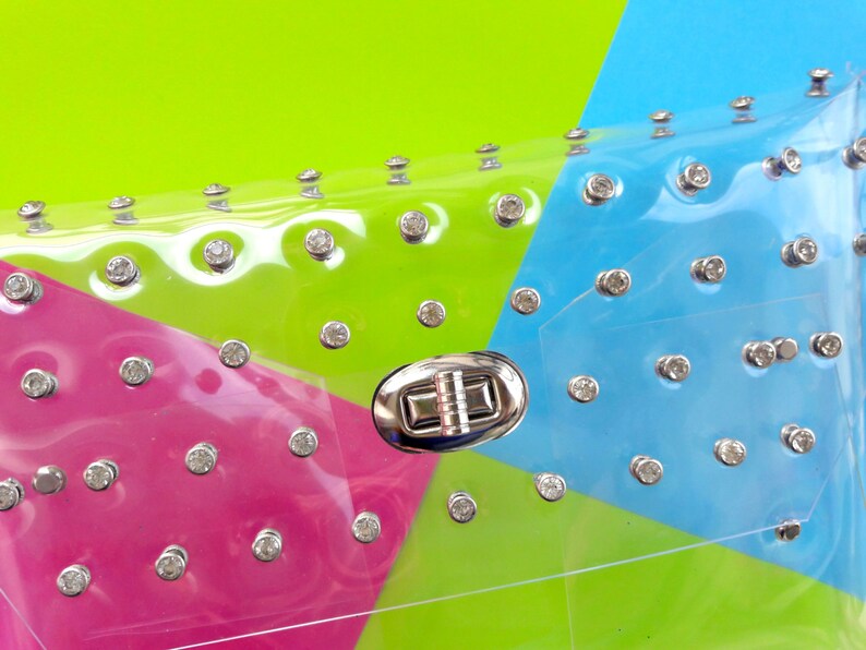 Puede incluir: Un bolso de mano transparente de pl&aacute;stico con un cierre plateado y pedrer&iacute;a. El bolso es rosa, verde y azul.