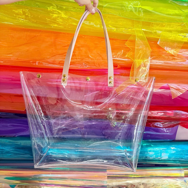 May include: A clear tote bag with light pink leather handles and gold-colored rivets. The bag is rectangular and transparent, allowing visibility of the contents. The background features a vibrant rainbow of colors.
