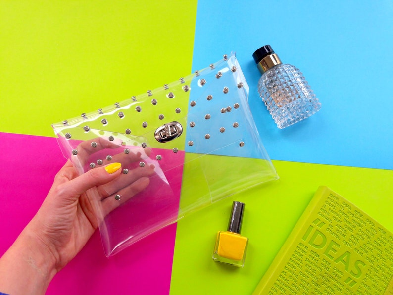 Puede incluir: Un bolso de mano transparente de pl&aacute;stico con tachuelas plateadas y un cierre plateado. El bolso se sostiene en una mano con esmalte de u&ntilde;as amarillo. El bolso est&aacute; sobre un fondo rosa, verde y azul brillante.