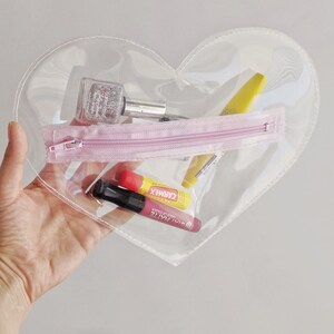 May include: A clear heart-shaped plastic zippered pouch with a pink zipper. The pouch contains a clear nail polish bottle with glitter, a pink lip gloss, a yellow lip balm tube with the text "CARMEX", and a red lip balm tube.