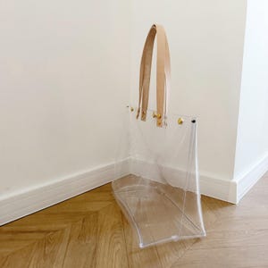 May include: A clear tote bag with tan leather handles and gold-colored rivets. The bag is made of transparent material, allowing visibility of the contents. The handles are attached with gold hardware. The bag is standing on a wooden floor.