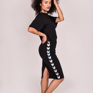 May include: A woman wearing a black t-shirt and a black skirt with a holographic chevron stripe down the side. She is wearing black sneakers and has blue lipstick.