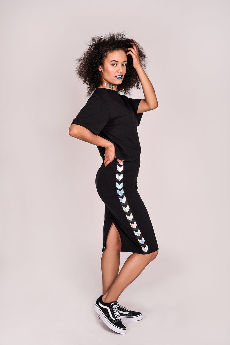 May include: A woman wearing a black t-shirt and a black skirt with a holographic chevron stripe down the side. She is wearing black sneakers and has blue lipstick.