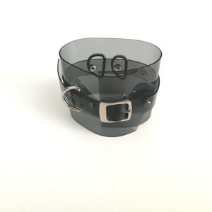 May include: A black translucent choker with a silver buckle and two silver rings. The choker is made of a flexible material and has a smooth finish.