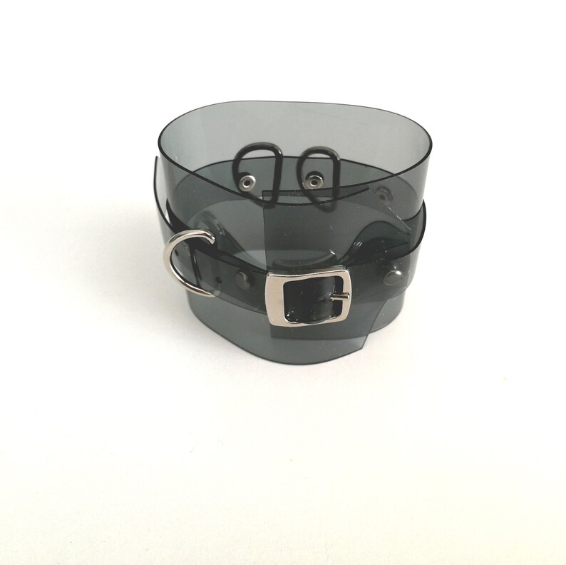 May include: A black translucent choker with a silver buckle and two silver rings. The choker is made of a flexible material and has a smooth finish.