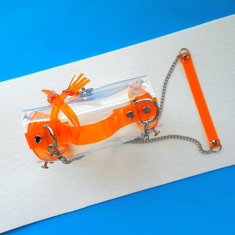 May include: A clear, rectangular bag with orange straps and accents. The bag features a silver chain and hardware. The straps are secured with rivets. The background is white and blue.