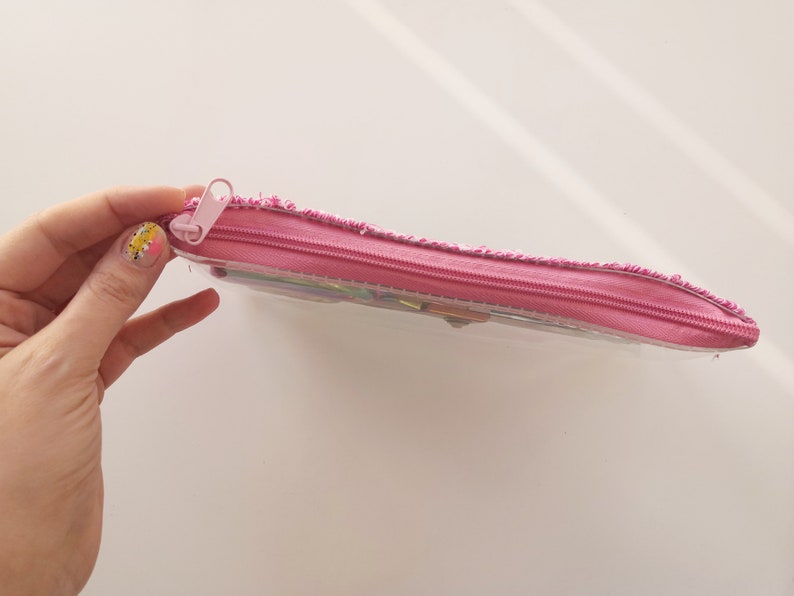 Pu&ograve; includere: Un astuccio rosa con un corpo in plastica trasparente e una cerniera rosa. L'astuccio &egrave; rettangolare e sembra essere parzialmente pieno. Il tirazip &egrave; anch'esso rosa.