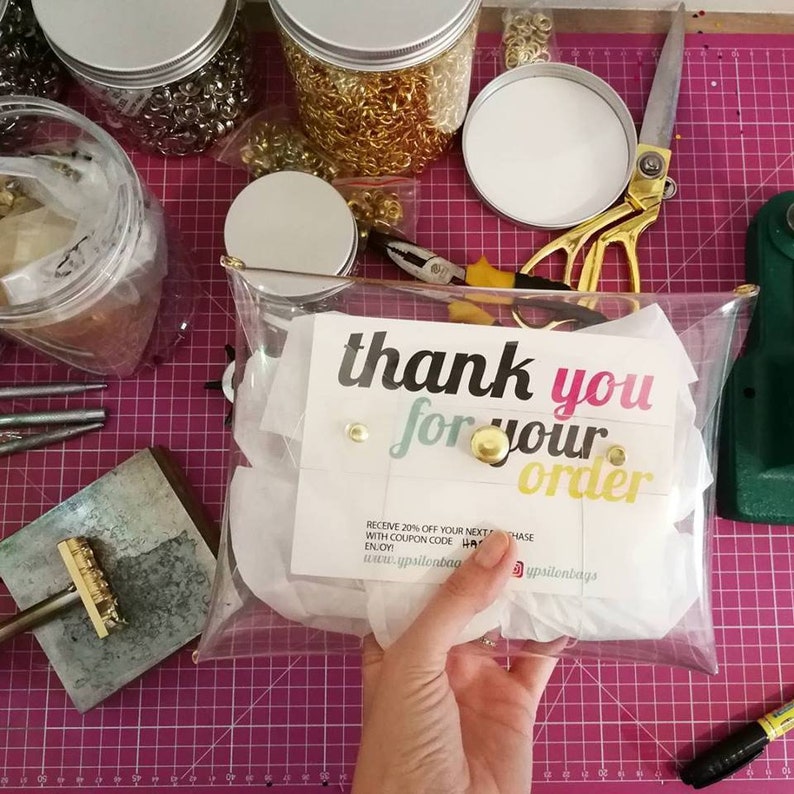 May include: A clear plastic bag with a thank you card inside. The card says "thank you for your order" and includes a discount code. The bag is sitting on a pink and white grid surface.