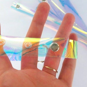 May include: A clear iridescent plastic strap with silver metal snaps. The strap is about 1 inch wide and has a holographic finish.