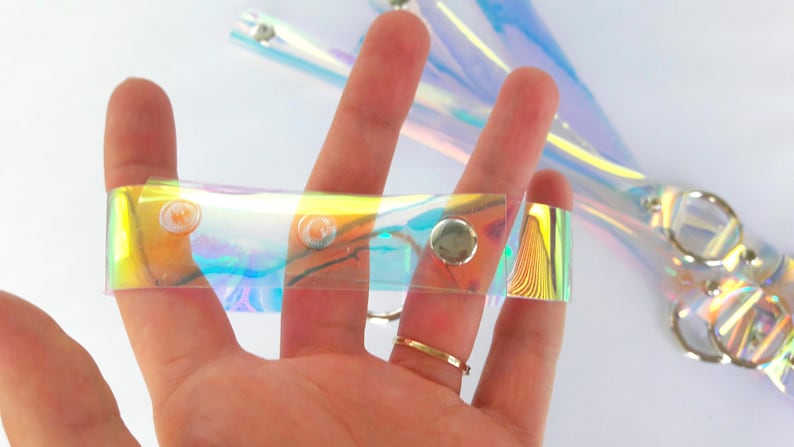 May include: A clear iridescent plastic strap with silver metal snaps. The strap is about 1 inch wide and has a holographic finish.