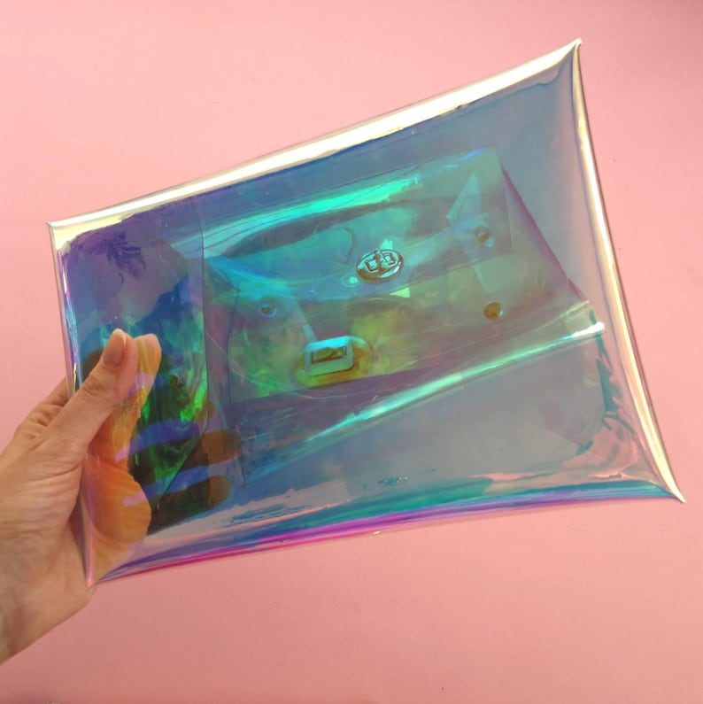 May include: A clear iridescent holographic clutch bag with a silver trim. The bag is made of a thin, flexible plastic material and has a zipper closure.