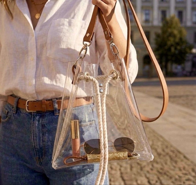 May include: A clear, triangular bucket bag featuring tan leather straps, silver-toned metal hardware, and a thick white rope drawstring closure. The transparent bag contains a gold-cased lipstick and a pair of dark-tinted sunglasses inside.
