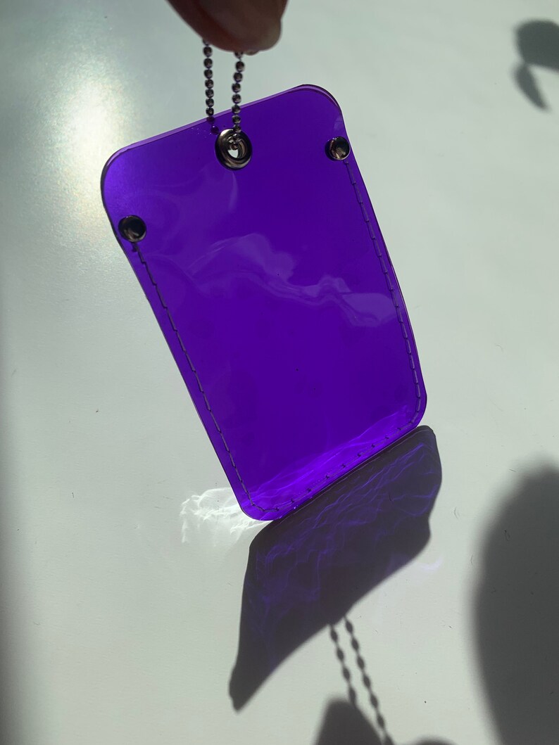 May include: A purple plastic keychain with a silver chain. The keychain is transparent and has a stitched border.