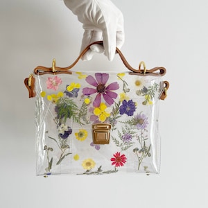 May include: A clear plastic handbag with a brown leather strap and gold hardware. The bag is decorated with pressed flowers in various colors, including purple, yellow, pink, and white.