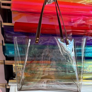 Puede incluir: Un bolso tote transparente e incoloro con dos correas de hombro de cuero marr&oacute;n oscuro sujetas con remaches de metal plateado. El bolso tiene un dise&ntilde;o rectangular de apertura superior con un compartimento principal y base plana.